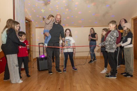 A group of people, including children, are in a room with wooden flooring and disco lights. A man holding a toddler stands next to a girl holding a red ribbon, while others watch and smile.