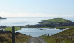 A gravel path leads downhill towards a coastal village with homes and green fields, flanked by a calm sea and distant islands under a clear blue sky.