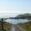 A gravel path leads downhill towards a coastal village with homes and green fields, flanked by a calm sea and distant islands under a clear blue sky.