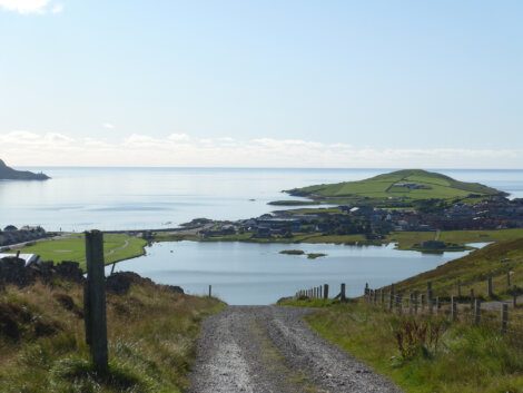 A gravel path leads downhill towards a coastal village with homes and green fields, flanked by a calm sea and distant islands under a clear blue sky.