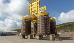 A large yellow offshore subsea structure with four cylindrical legs marked with depth measurements, situated outdoors on a concrete surface against a backdrop of hills and cloudy sky.