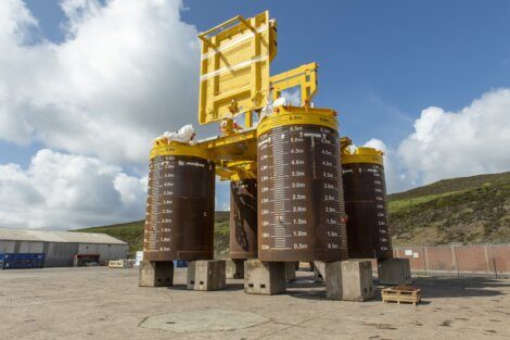 A large yellow offshore subsea structure with four cylindrical legs marked with depth measurements, situated outdoors on a concrete surface against a backdrop of hills and cloudy sky.