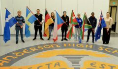 Seven students stand in a line holding various national flags on a floor with "Anderson High School" written on it.