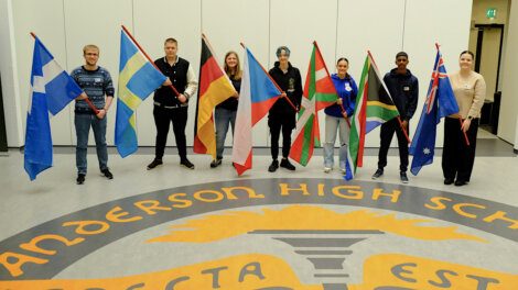 Seven students stand in a line holding various national flags on a floor with "Anderson High School" written on it.