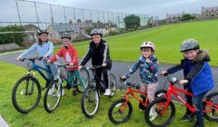 Five children in helmets stand with their bicycles on a paved path next to a grassy field and fence, with houses visible in the background on a cloudy day.