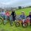 Five children in helmets stand with their bicycles on a paved path next to a grassy field and fence, with houses visible in the background on a cloudy day.