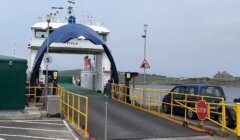 Ferry docked at port with ramp extended, surrounded by yellow railings and traffic signs. Cars and a building are visible in the background.