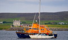 An orange lifeboat assists a white sailboat near a coastal shoreline with houses in the background.