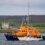 An orange lifeboat assists a white sailboat near a coastal shoreline with houses in the background.