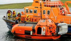 Lifeboat crew in yellow suits conducts a drill or rescue operation on an orange rescue boat near a green coastal area. The boat is marked "Lifeboats" and "PULP MILL–JOE BIRCHARD.