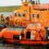 Lifeboat crew in yellow suits conducts a drill or rescue operation on an orange rescue boat near a green coastal area. The boat is marked "Lifeboats" and "PULP MILL–JOE BIRCHARD.