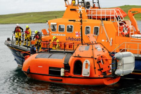 Lifeboat crew in yellow suits conducts a drill or rescue operation on an orange rescue boat near a green coastal area. The boat is marked "Lifeboats" and "PULP MILL–JOE BIRCHARD.