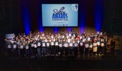 Large group of award recipients on stage holding certificates at the Saltire Awards Ceremony 2024. A projector screen behind them displays the event's logo and name.