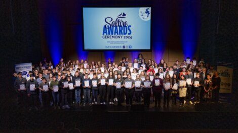 Large group of award recipients on stage holding certificates at the Saltire Awards Ceremony 2024. A projector screen behind them displays the event's logo and name.