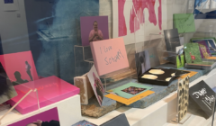 An art display featuring various colorful prints, books, and postcards arranged on shelves and stands, seen through a reflective glass.