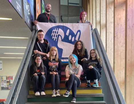 A group of eight people are seated and standing on a staircase holding a large banner with a cartoonish creature and text. They are indoors, and some smile at the camera.