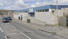 A white and blue building with a sign reading "Shetland Recreational Trust, Shetland Bowls Centre" on a partially cloudy day. Cars are driving on the street in front of it.