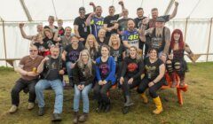 A group of people, men and women, of various ages, some with children, pose enthusiastically inside a tent. They are casually dressed, and a few wear graphic tees and biker gear.