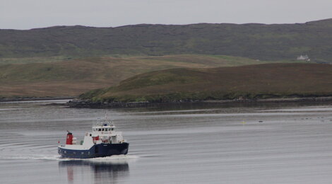 A small ship sails through calm waters with hills and a few buildings visible in the background.