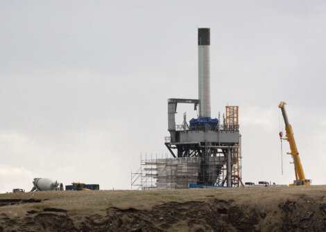 A construction site featuring a tall industrial structure with scaffolding, a crane, various construction materials, and vehicles on a barren landscape under a cloudy sky.