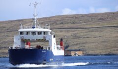 A blue and white ferry named "Bíggja" sails on calm waters with a hilly, grass-covered shoreline and a small structure in the background.