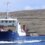 A blue and white ferry named "Bíggja" sails on calm waters with a hilly, grass-covered shoreline and a small structure in the background.