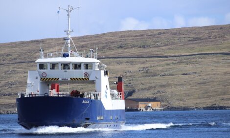 A blue and white ferry named "Bíggja" sails on calm waters with a hilly, grass-covered shoreline and a small structure in the background.