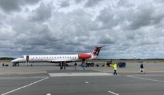 An airplane with red and white tail livery is parked on the tarmac under a cloudy sky. Ground crew and passengers are visible near the aircraft, and various baggage carts are in the background.