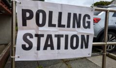 A white banner with bold black text reads "POLLING STATION" attached to a metal fence outside a building. A parked car is visible in the background.