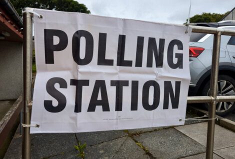 A white banner with bold black text reads "POLLING STATION" attached to a metal fence outside a building. A parked car is visible in the background.