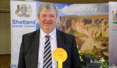 A man in a suit and striped tie stands in front of a backdrop featuring "Orkney" and "Shetland Islands Council" logos, holding a large yellow rosette.