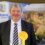 A man in a suit and striped tie stands in front of a backdrop featuring "Orkney" and "Shetland Islands Council" logos, holding a large yellow rosette.