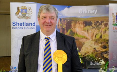 A man in a suit and striped tie stands in front of a backdrop featuring "Orkney" and "Shetland Islands Council" logos, holding a large yellow rosette.