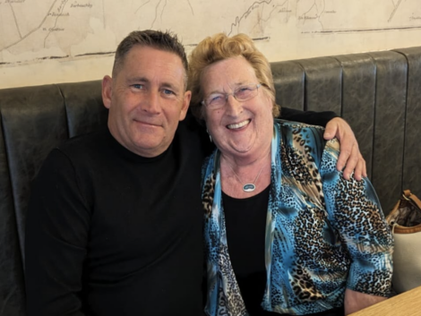 A man in a black shirt and an older woman in a blue patterned blouse are sitting together in a booth, smiling at the camera. The woman has her arm around the man's shoulders.