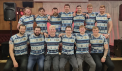 A group of fifteen men wearing matching festive-patterned shirts, posing together inside a room with red and white decorations.