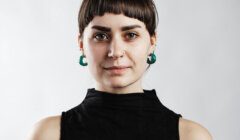 Person with short, dark hair and green earrings wearing a black sleeveless top, facing the camera against a plain white background.