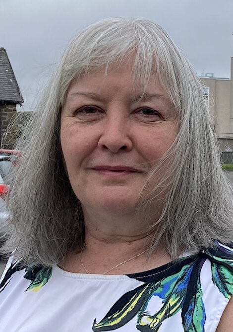 Woman with gray hair and a thoughtful expression, wearing a white top with colorful patterns, standing outdoors with buildings and vehicles in the background.