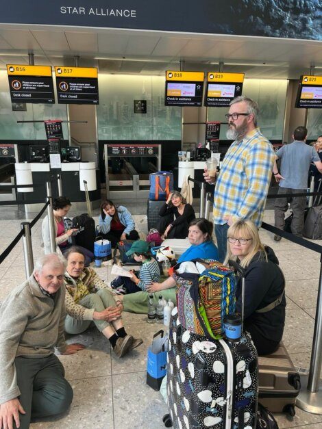 A group of people sits on the floor near luggage and check-in counters at an airport, appearing to wait. The check-in counters are for B16, B17, and B19, belonging to Star Alliance.