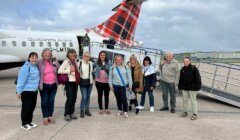 Nine people stand in front of a small plane with Scotland's Airline branding, smiling at the camera. The plane is on a tarmac, with a staircase leading to the entrance. The weather is cloudy.