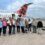 Nine people stand in front of a small plane with Scotland's Airline branding, smiling at the camera. The plane is on a tarmac, with a staircase leading to the entrance. The weather is cloudy.