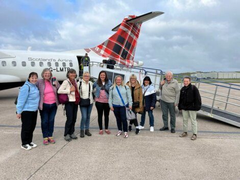 Nine people stand in front of a small plane with Scotland's Airline branding, smiling at the camera. The plane is on a tarmac, with a staircase leading to the entrance. The weather is cloudy.