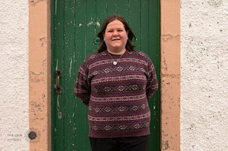 A person with shoulder-length hair stands in front of a green door, wearing a patterned sweater and dark pants. They are facing the camera with their arms behind their back.