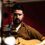 A man with a beard and brown shirt plays an acoustic guitar indoors with a focused expression.