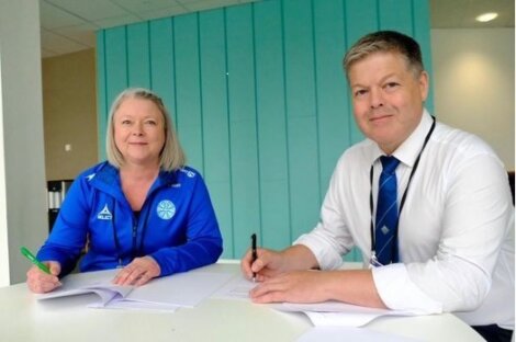 Two people sit at a table signing documents. One is a woman in a blue sports jacket, and the other is a man in a white shirt and blue tie. Both are holding pens and facing the camera.