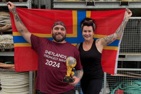 A man holds a trophy while wearing a "Shetlands Strongest Man 2024" shirt and standing next to a woman. Both are smiling, and a flag is held behind them.