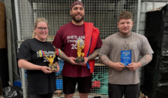 Three people pose indoors holding trophies, each wearing shirts with "Shetland Strongest 2023" text. The man in the middle also holds a flag draped over his shoulder.