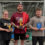 Three people pose indoors holding trophies, each wearing shirts with "Shetland Strongest 2023" text. The man in the middle also holds a flag draped over his shoulder.