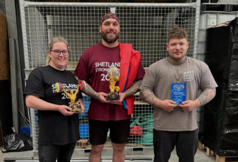 Three people pose indoors holding trophies, each wearing shirts with "Shetland Strongest 2023" text. The man in the middle also holds a flag draped over his shoulder.