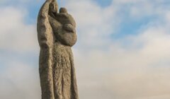 A stone statue depicts a veiled woman holding a child against a backdrop of a cloudy sky.