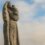 A stone statue depicts a veiled woman holding a child against a backdrop of a cloudy sky.